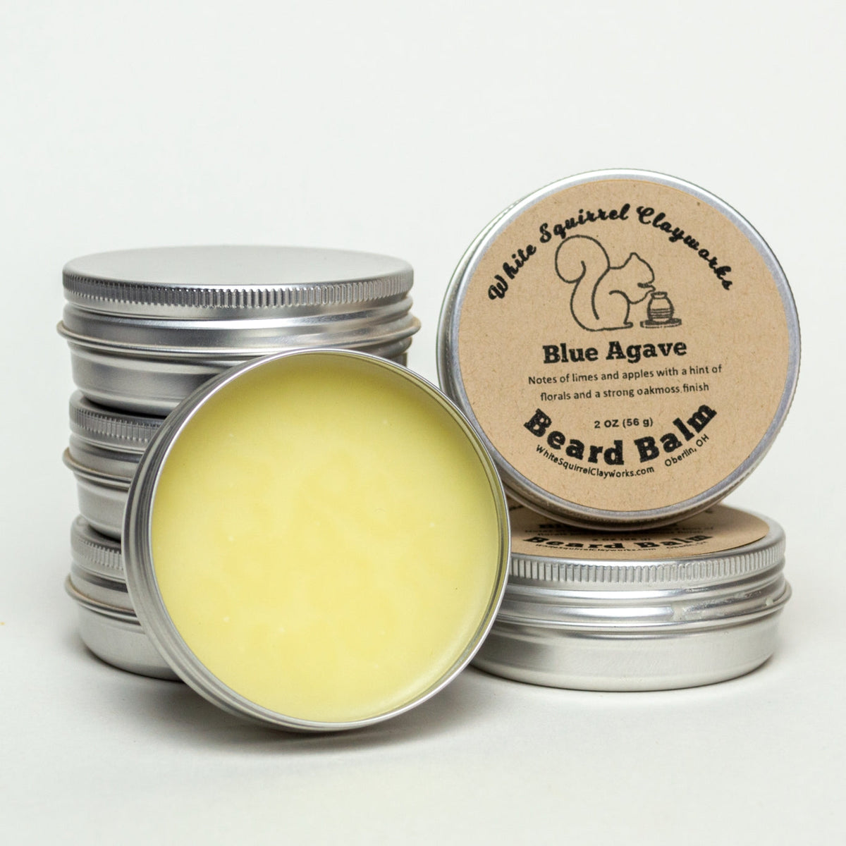 Stack of metal beard balm tins, one open to reveal pale yellow balm; label reads Blue Agave Beard Balm by White Squirrel Clayworks.