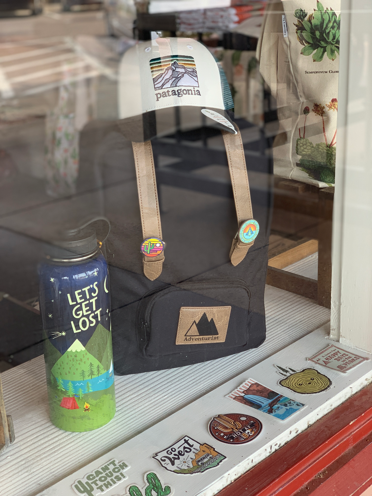 Patagonia cap perched on a black backpack with tan straps in a storefront display, alongside a water bottle and a plant print tote.