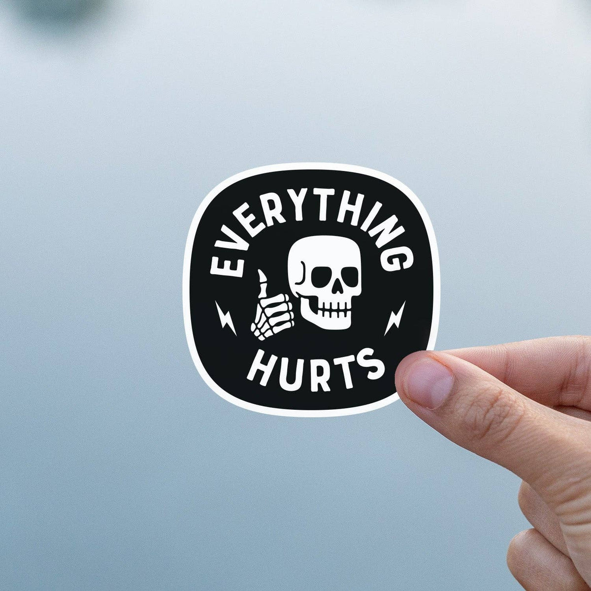 Everything Hurts sticker held against a pale blue gray background with the skull and bold text.