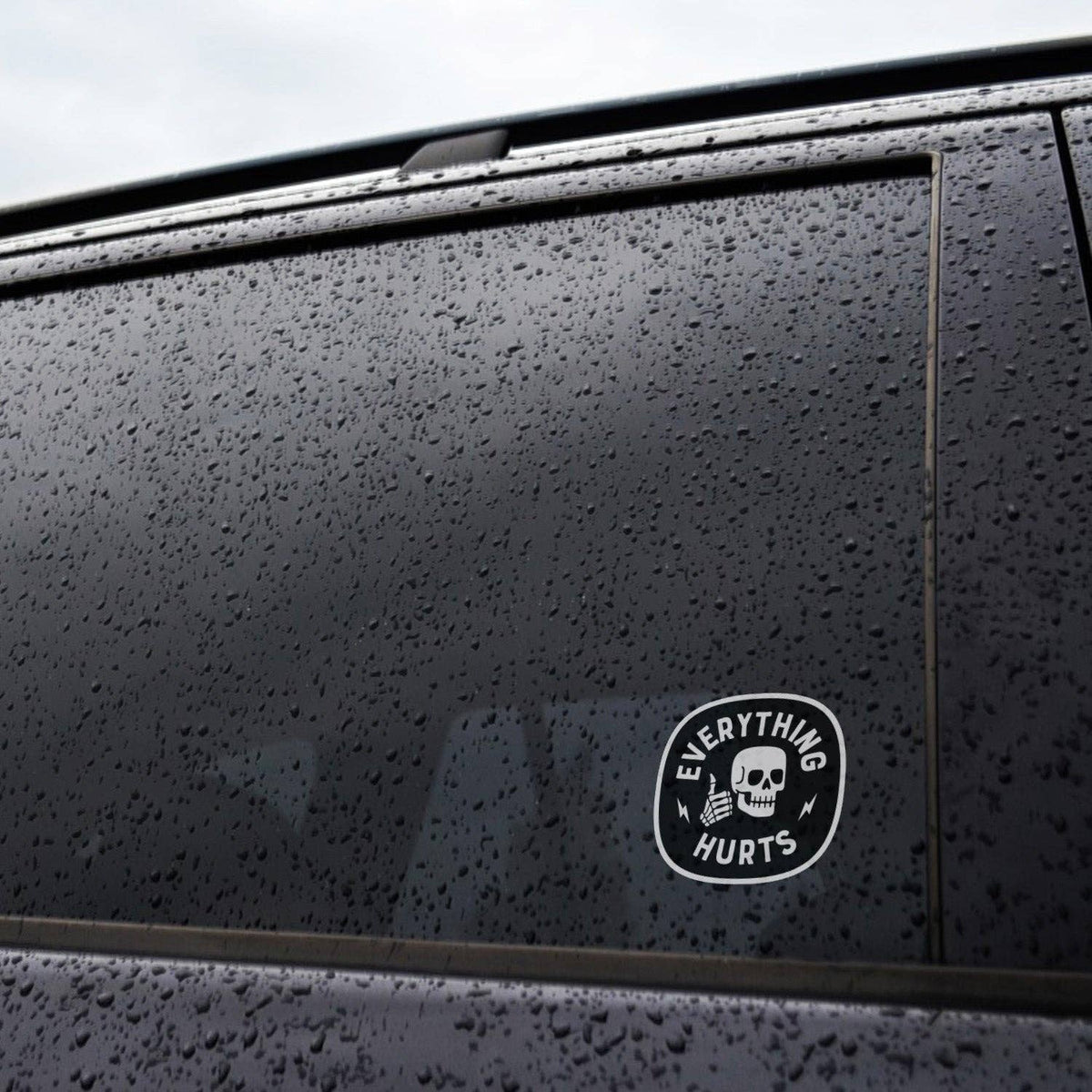 Rainy car window with numerous droplets and a small Everything Hurts sticker visible on the glass