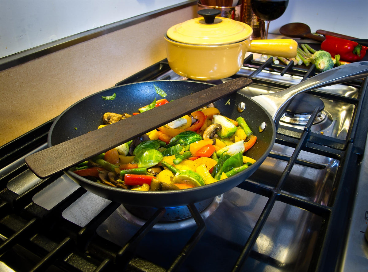 Wide flat wooden saute spatula across a pan of sautéed vegetables on a gas stove.