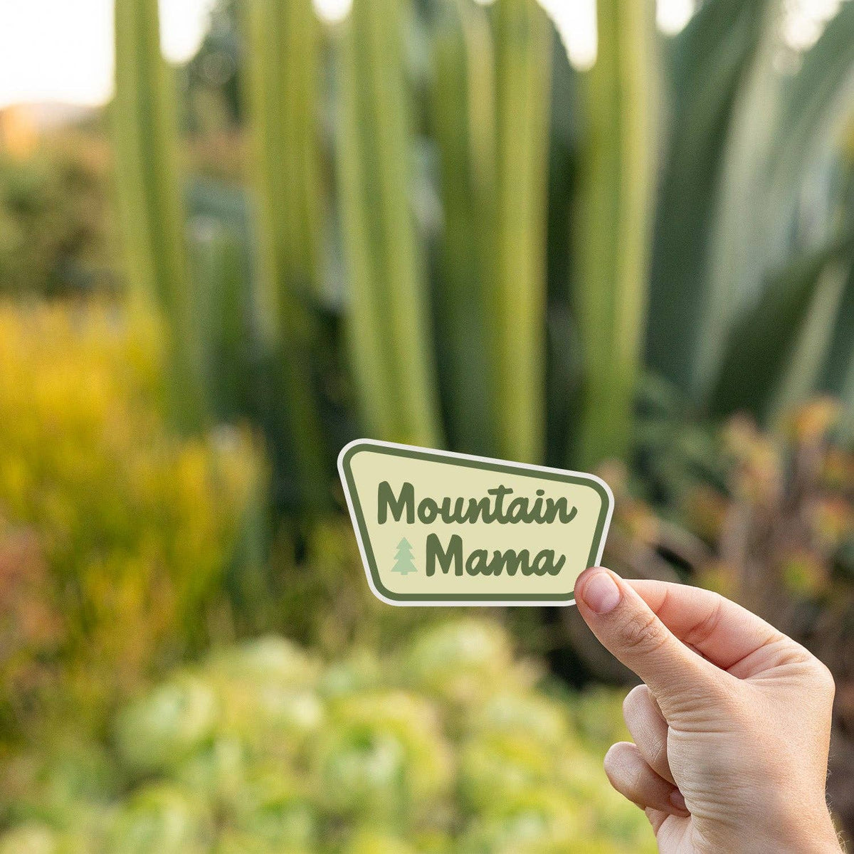 Hand holding Mountain Mama sticker outdoors with tall green plants in the background