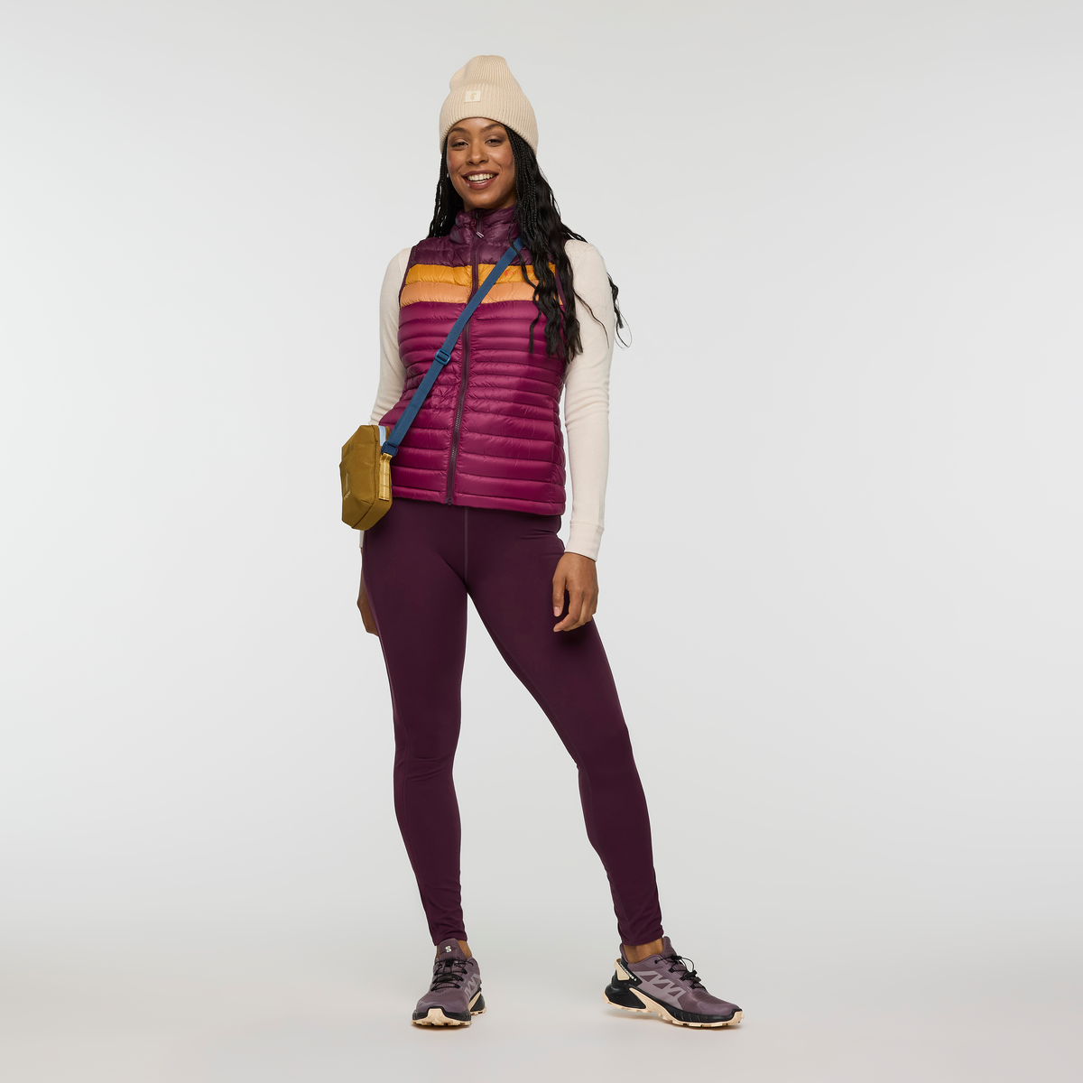 Front view of a woman wearing Cotopaxi Fuego Down Vest in purple with orange chest stripe, beige hat, and crossbody bag.