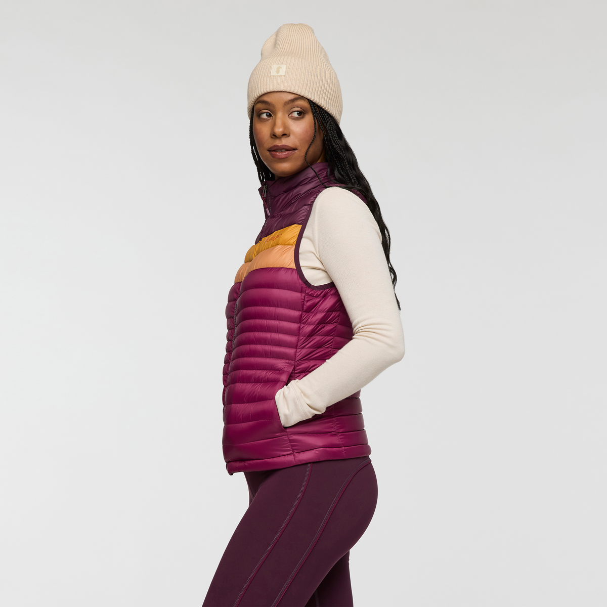Side profile of a woman wearing Cotopaxi Fuego Down Vest in purple with amber chest stripe, beige sleeves.