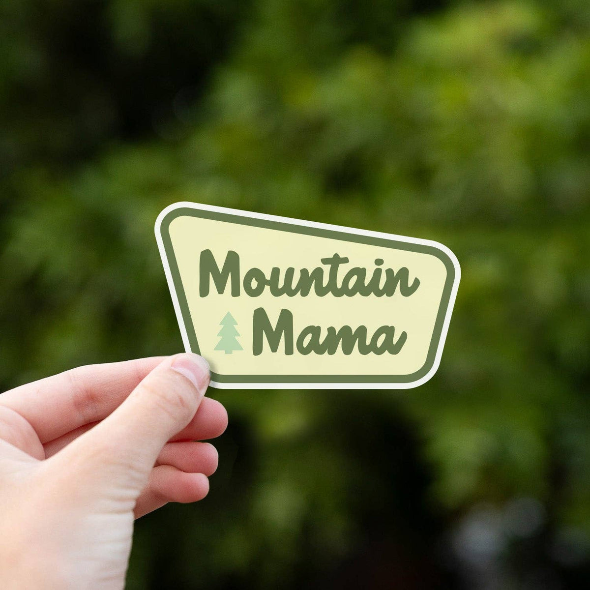 Hand holding a Mountain Mama sticker with a pale yellow label and pine tree icon against a blurred green background.