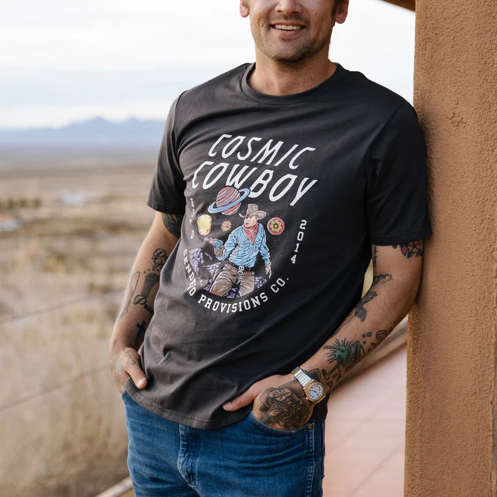 Man in Cosmic Cowboy T-Shirt leaning against a stucco wall with a desert landscape behind