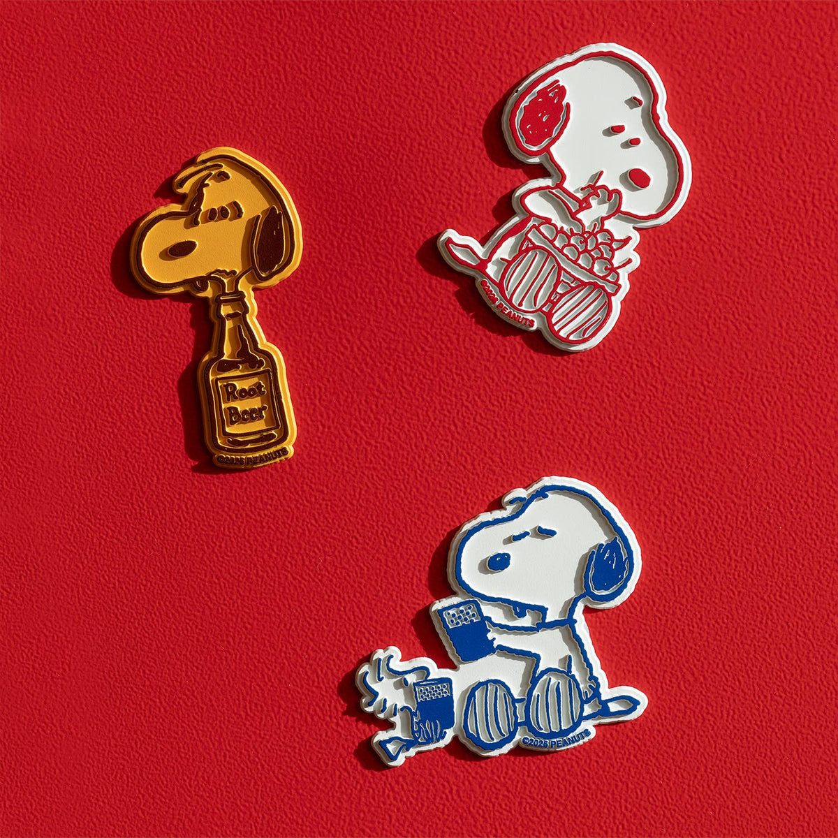Three Peanuts Snoopy magnets on a red surface including a yellow Snoopy with a root beer bottle and two white Snoopy poses