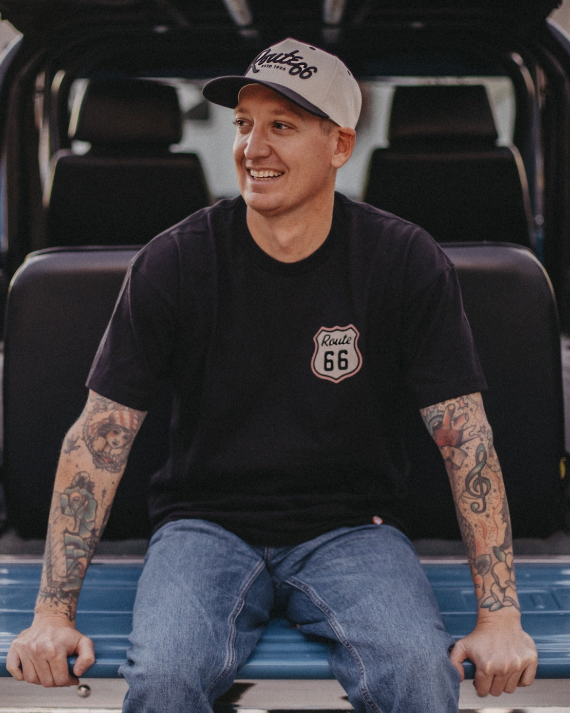 Smiling man in black Route 66 tee and beige cap sitting on a car tailgate