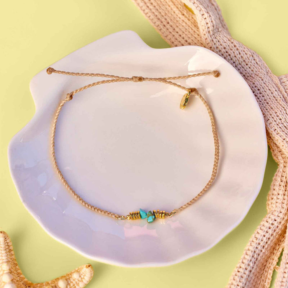 Beige braided anklet with turquoise chips and tiny gold beads on a white plate.