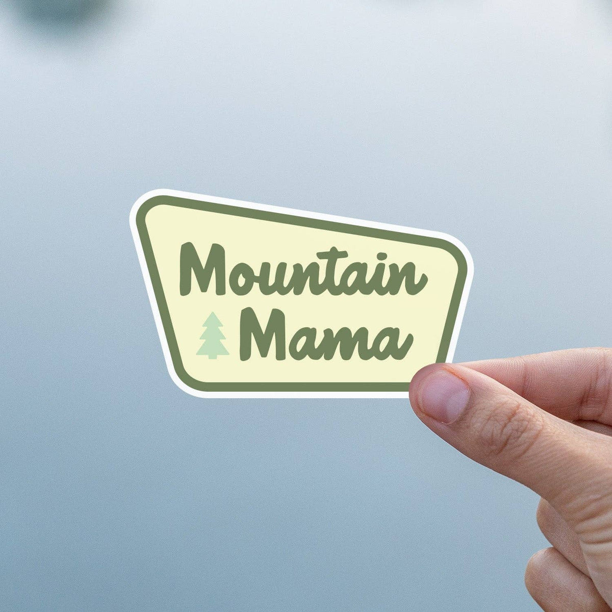 Mountain Mama sticker featuring a pine tree icon held by fingers against a pale blue background