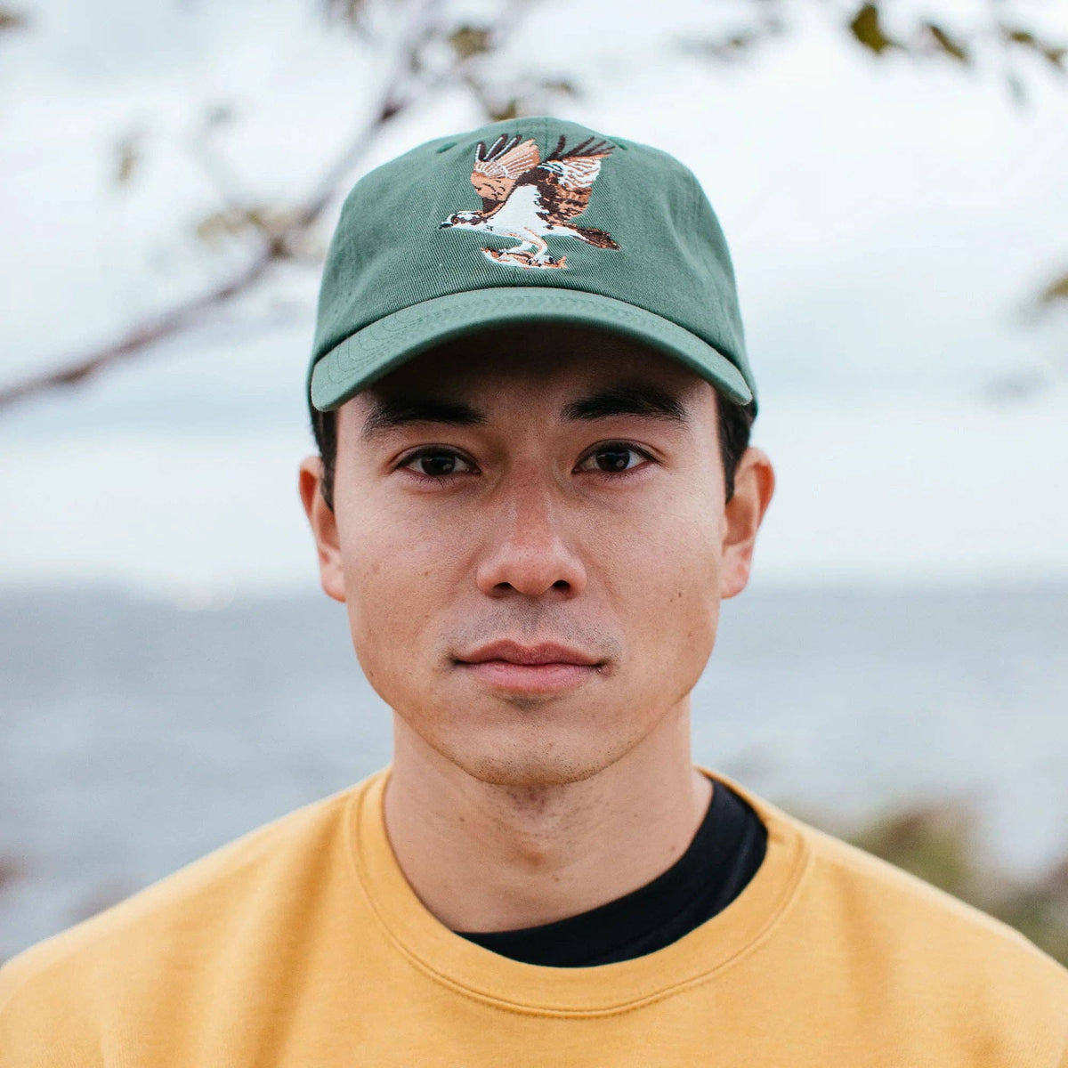 Front view of a green cap with osprey embroidery worn by a person in a tan shirt