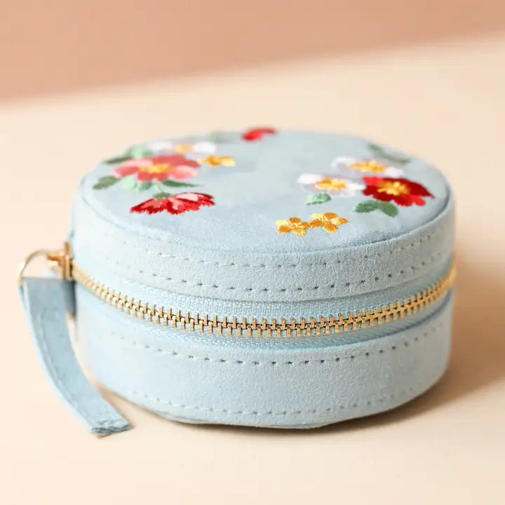 Light blue round velvet jewelry case with colorful floral embroidery on the lid and a gold zipper.