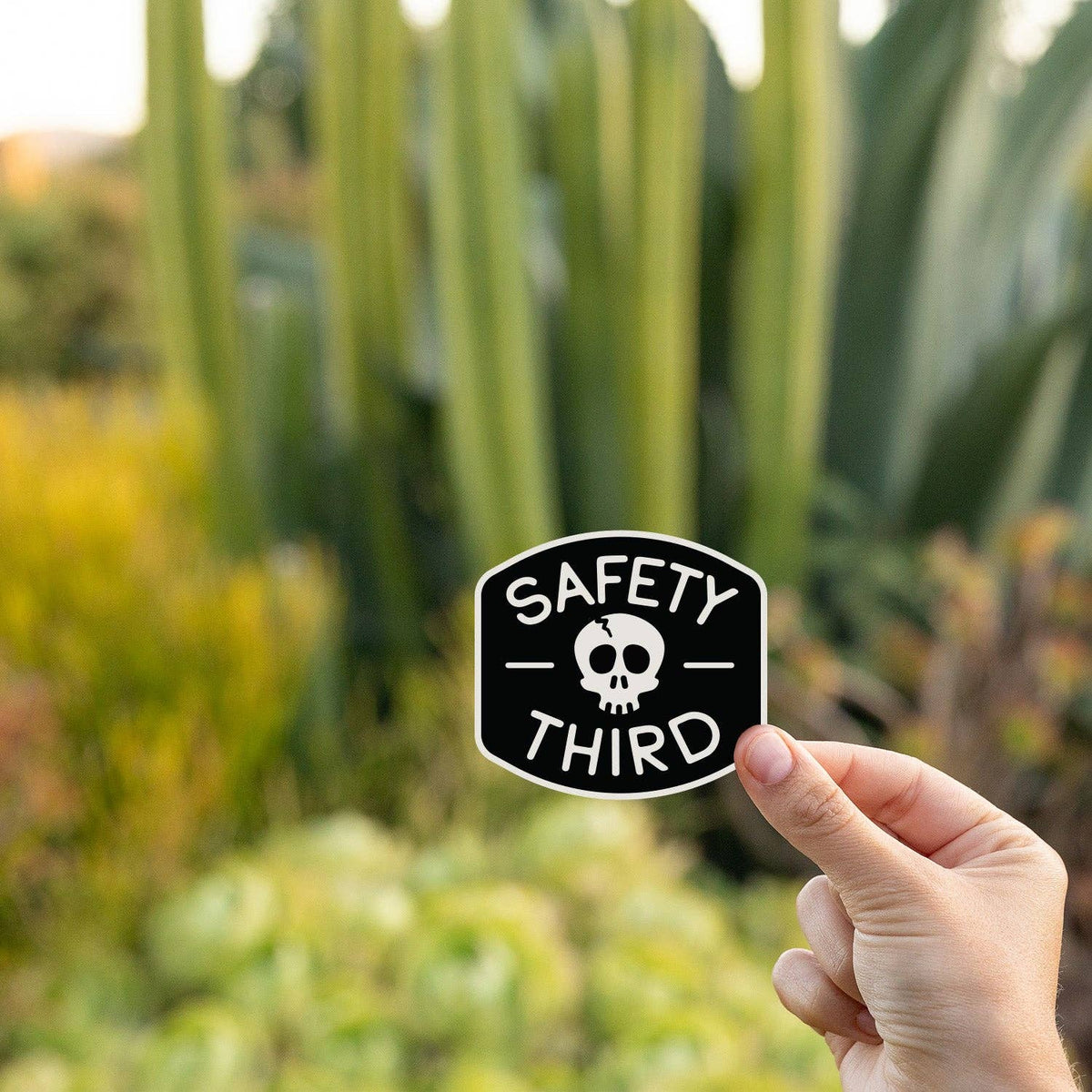 Black shield shaped Safety Third sticker with a white skull held by fingers against a blurred outdoor background