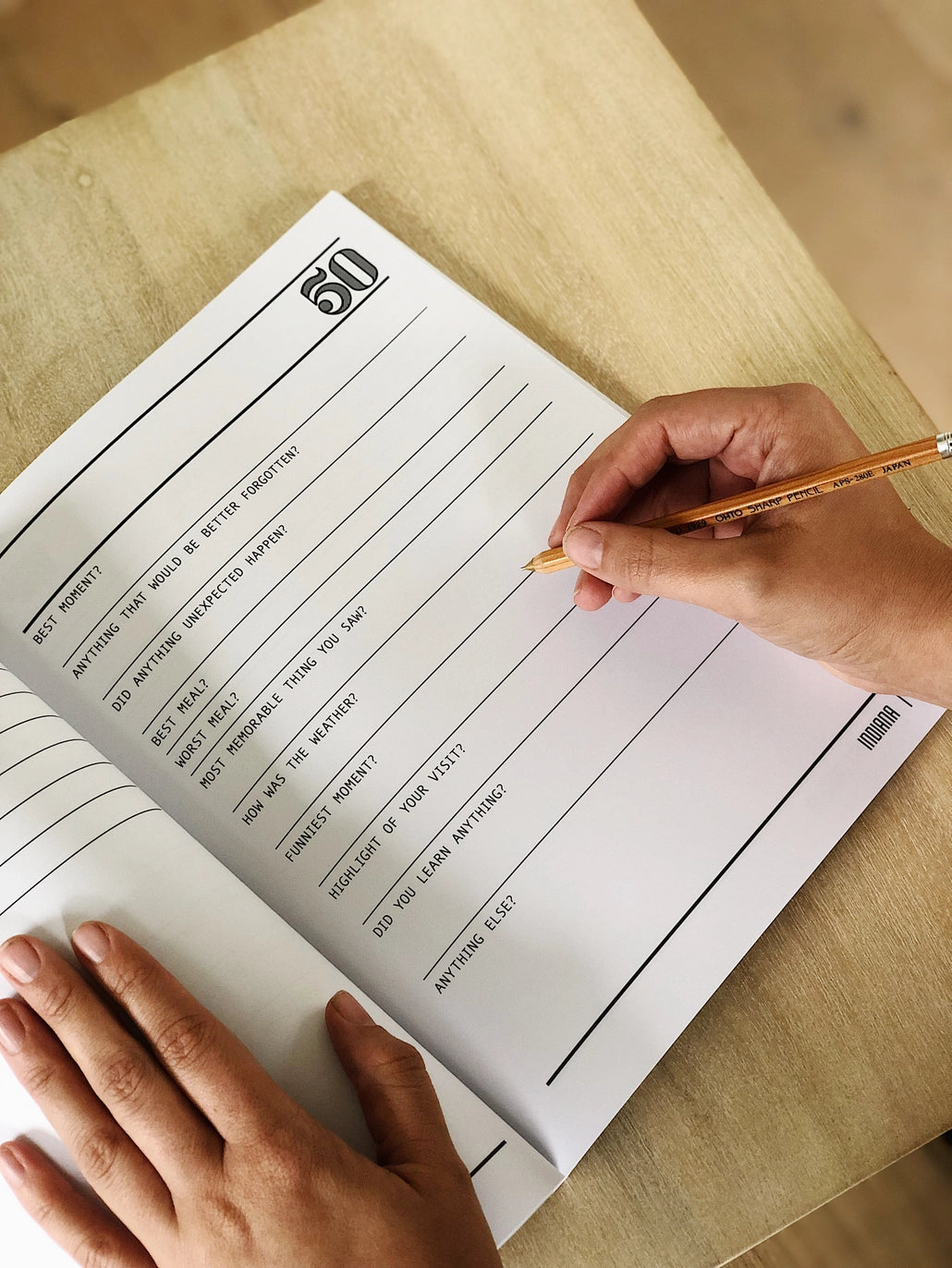 Hands write in a travel journal page filled with prompts and a 50 emblem at the top.