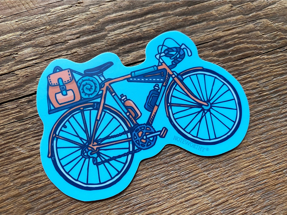 Blue bicycle sticker with brown pannier bag and gear on a wooden surface