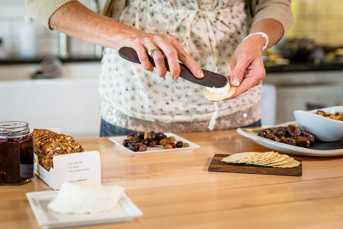 Hands using a wooden spreader to spread a creamy topping on a cracker