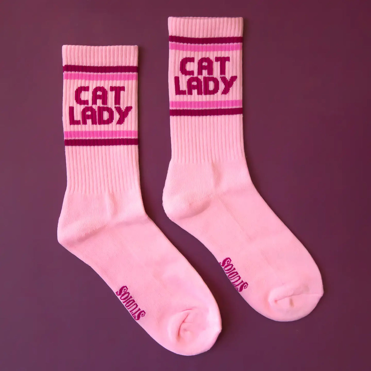 Two pink ribbed crew socks with CAT LADY text and bold striped bands against a mauve backdrop.
