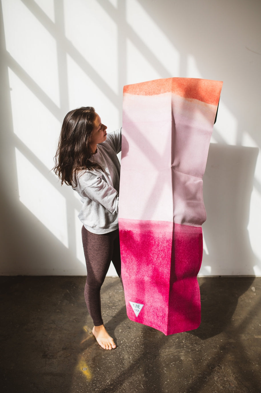 Woman barefoot indoors holds a tall gradient suede yoga travel mat with pink and peach tones.