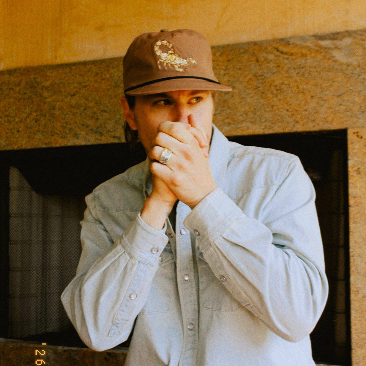Close-up of a person wearing a brown Scorpion Hat with embroidered scorpion and hands near their mouth