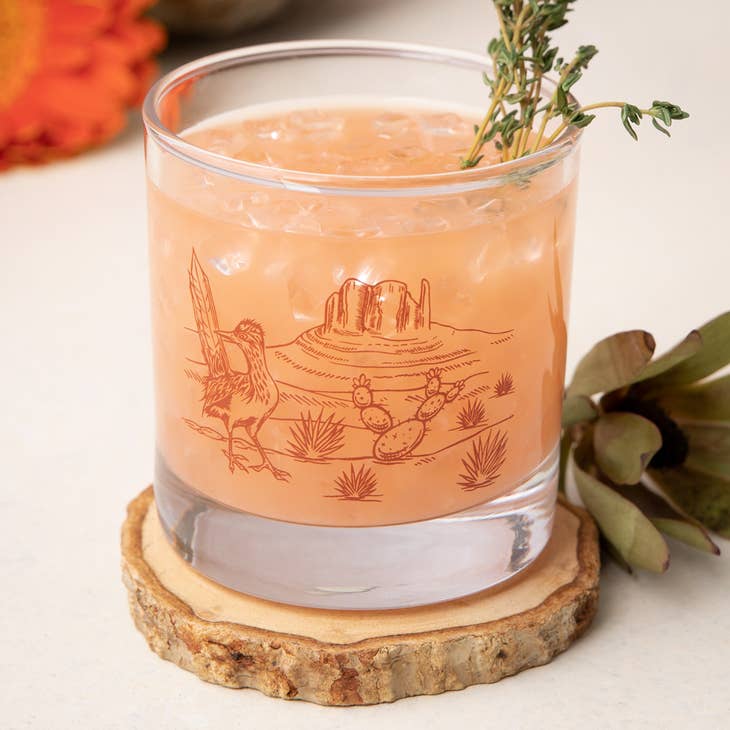 Desert Rocks Glass filled with a peach cocktail, ice and a thyme sprig garnish, desert scene etched on the glass