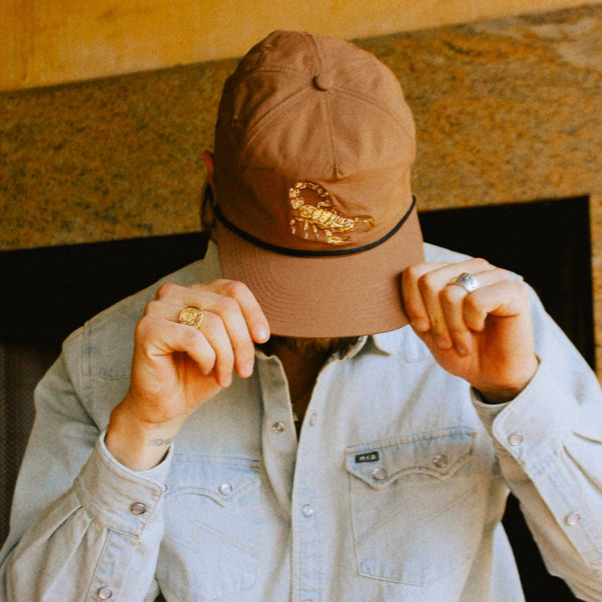 Beige cap with a gold scorpion emblem worn by a person adjusting the brim over a denim shirt.