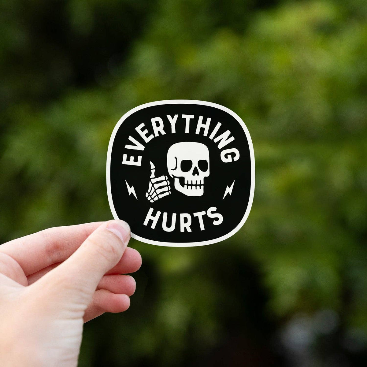 Hand holding an Everything Hurts sticker with skull image against a blurred green outdoor background