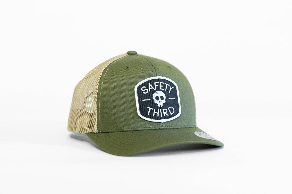 Three quarter view of a green front panel trucker hat with tan mesh and a front patch reading Safety Third with skull emblem.