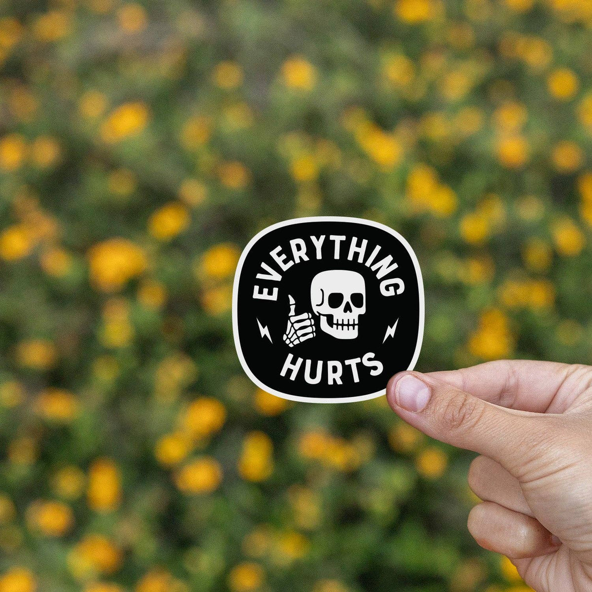 Everything Hurts sticker held in front of a field of yellow flowers with the skull logo.