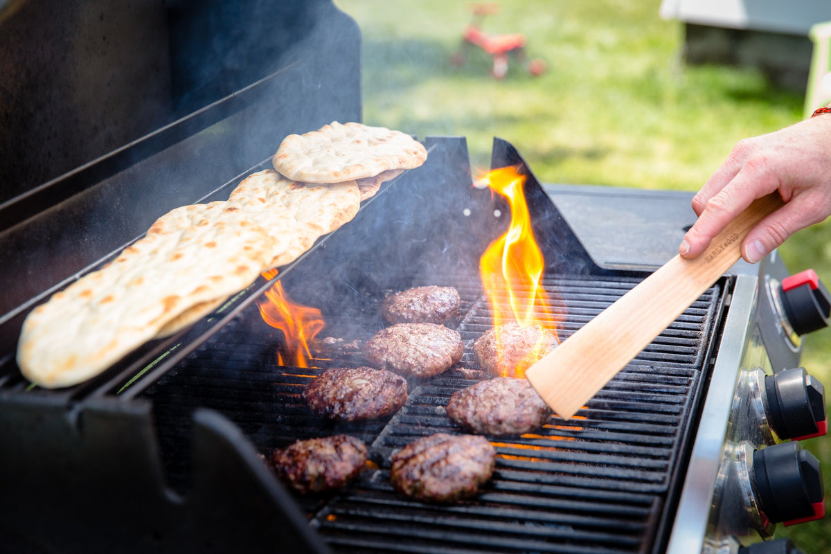 Large flat wooden saute spatula resting across a skillet on a barbecue grill with colorful vegetables.