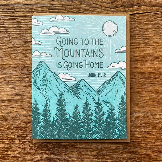 Going to the Mountains greeting card on a wood surface with teal mountains and sun illustration