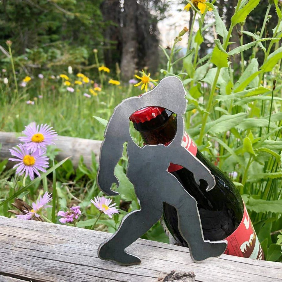 Bigfoot shaped metal bottle opener set on a weathered wooden railing with wildflowers; bottle neck visible through the torso opening.