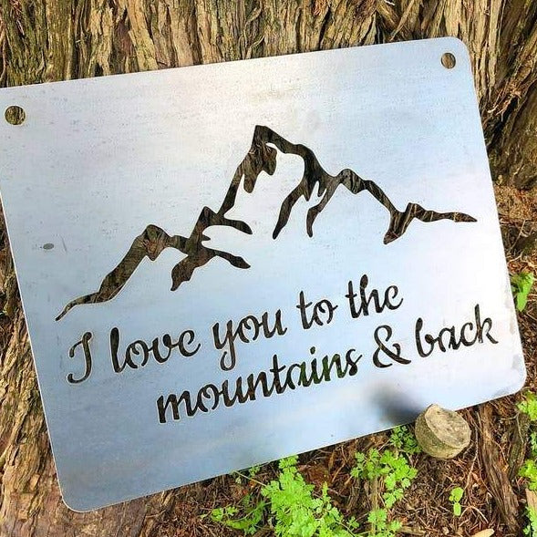 Large steel sign with a mountain silhouette and the words I love you to the mountains & back leaning against a tree.