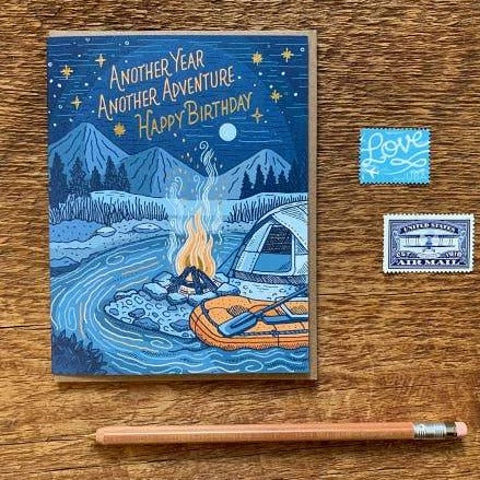 Campfire Birthday Card featuring a blue night campsite with mountains, campfire, tent and inflatable orange raft, shown with pencil and stamps.