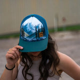 Misty Mountains unisex trucker hat worn by a person with teal brim and blue mesh and a mountain landscape patch