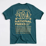 Back view of teal National Parks Travel Club T shirt with a cream circular logo featuring trees and sun