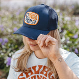 Navy curved trucker hat with an orange edged patch displaying the Savage Sands logo and desert illustration