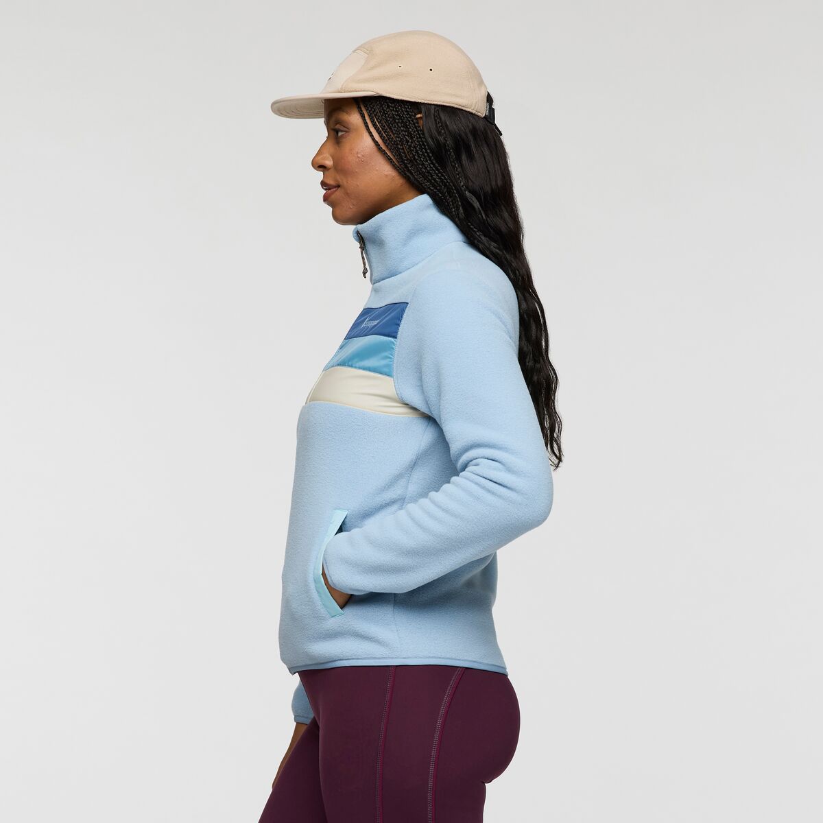 Profile view of a woman in a light blue Cotopaxi fleece jacket with a zip collar and color block chest stripes, wearing a beige cap