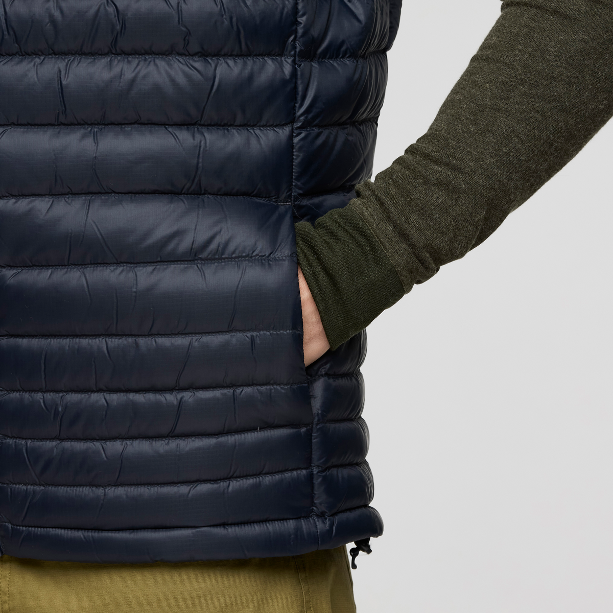 Navy Cotopaxi Fuego Down Vest with horizontal quilting and a side pocket against a dark green sleeve.