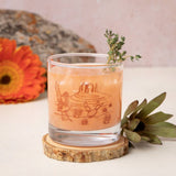 Desert Rocks Glass with canyon and cactus scene etched on the glass, peach cocktail and thyme garnish on a wooden coaster