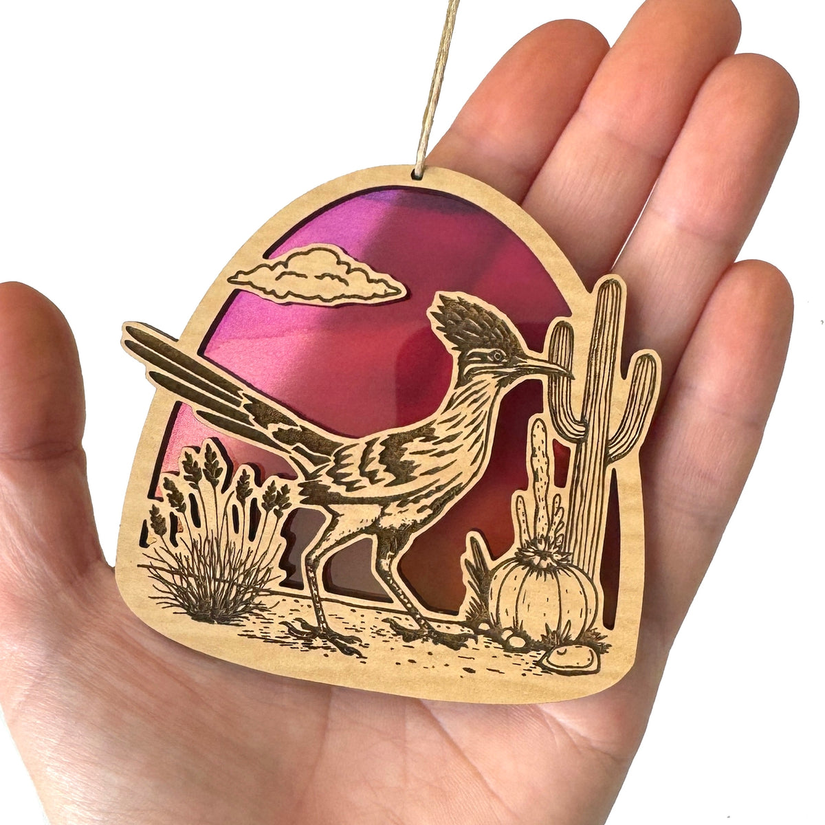 Front view of a desert roadrunner ornament with pink purple sunset and cactus, held in a hand