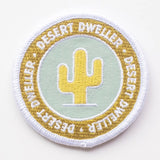 Desert Dweller patch showing a yellow cactus on a pale blue circle with white and gold border