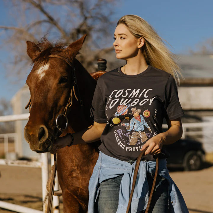 Woman wearing Cosmic Cowboy T-Shirt beside a brown horse in a sunny outdoor setting