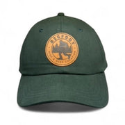 A green adjustable dad hat with a woven patch on the front featuring an image of a mountain and the text 'Bigfoot Hide & Seek World Champ'. It has a brass buckle for adjustability.