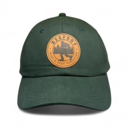 A green adjustable dad hat with a woven patch on the front featuring an image of a mountain and the text 'Bigfoot Hide & Seek World Champ'. It has a brass buckle for adjustability.
