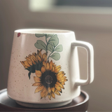 Sunflower illustration on a white stoneware mug with a curved handle and soft lighting.