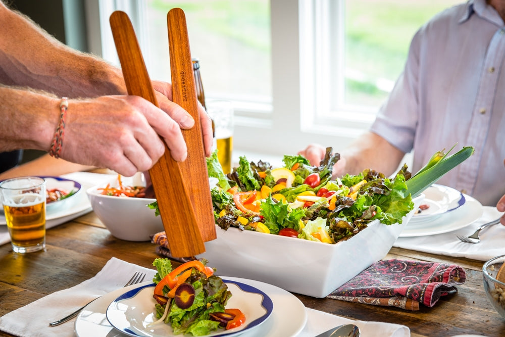 Large flat wooden saute spatula used to serve a colorful salad from a white dish.