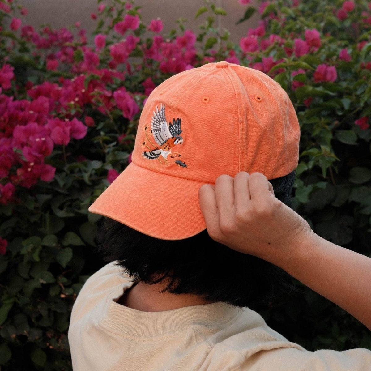 Side view of a person wearing an orange American Kestrel Washed Dad Hat with bird embroidery and pink flowers in the background