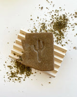 Brown desert rain soap bar with a fork imprint surrounded by dried herbs on a white surface.