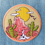 Circular patch with a desert scene of red rock formations, green cacti, white clouds and a yellow sun