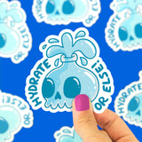 Close up of a light blue skull sticker with a white outline on a bright blue background being peeled by a finger